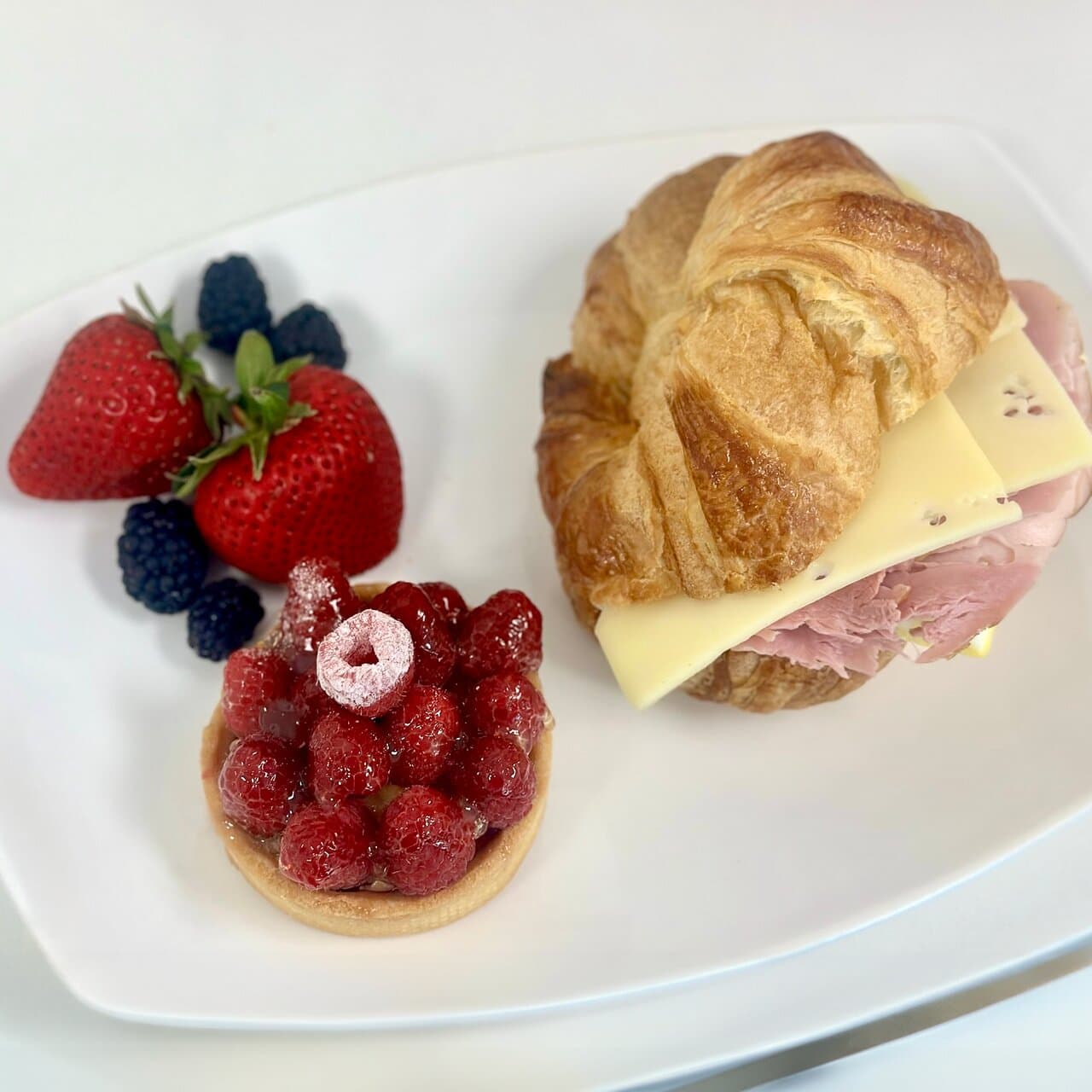 A ham and cheese croissant sandwich plated with a raspberry tart and fresh berries